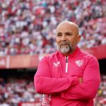 Jorge Sampaoli, entrenador de fútbol con barba canosa y chaqueta deportiva rosa del Sevilla FC, con los brazos cruzados y expresión seria frente a una grada llena de aficionados.