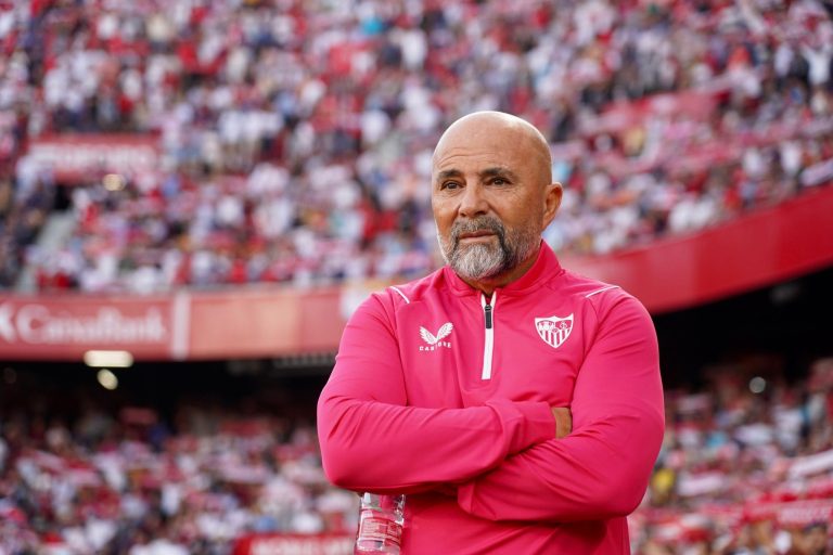 Jorge Sampaoli, entrenador de fútbol con barba canosa y chaqueta deportiva rosa del Sevilla FC, con los brazos cruzados y expresión seria frente a una grada llena de aficionados.