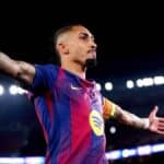 Raphinha, jugador del FC Barcelona con la camiseta azulgrana y brazalete de capitán, celebrando con los brazos extendidos y gesto desafiante en un estadio iluminado.