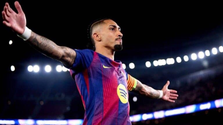 Raphinha, jugador del FC Barcelona con la camiseta azulgrana y brazalete de capitán, celebrando con los brazos extendidos y gesto desafiante en un estadio iluminado.
