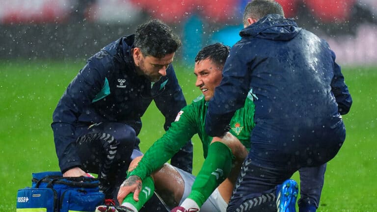 Jugador de fútbol recibiendo asistencia médica de dos fisioterapeutas en el césped durante un partido, práctica que la FIFA quiere limitar con una nueva norma que obligará a permanecer un minuto fuera del campo.