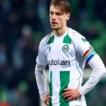 Stije Resink, joven futbolista neerlandés, con la equipación blanca y verde del Groningen, de pie en el campo con las manos en las caderas y expresión concentrada.