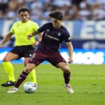 Acción de juego entre un futbolista del Eibar controlando el balón y un árbitro siguiendo la jugada en Ipurúa.