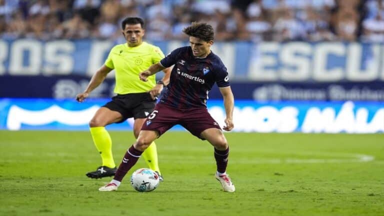 Acción de juego entre un futbolista del Eibar controlando el balón y un árbitro siguiendo la jugada en Ipurúa.