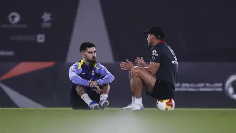 Diego Simeone sentado sobre un balón conversando intensamente con un jugador del Atlético de Madrid durante un entrenamiento nocturno.