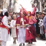 Multitud de personas celebrando el Carnaval de Tolosa en una calle estrecha; en el centro destaca un hombre disfrazado de obispo con mitra roja y capa, rodeado de gente con boinas rojas y palos de madera.