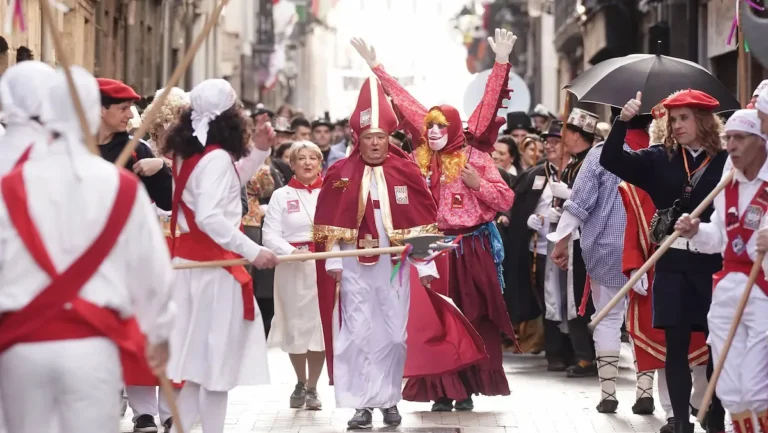 Multitud de personas celebrando el Carnaval de Tolosa en una calle estrecha; en el centro destaca un hombre disfrazado de obispo con mitra roja y capa, rodeado de gente con boinas rojas y palos de madera.