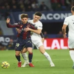 Acción del derbi valenciano donde un jugador del Valencia CF con equipación blanca sujeta por la cintura a un jugador del Levante UD con camiseta azulgrana, mientras el balón rueda cerca de ellos en el césped.