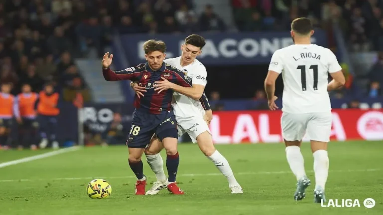 Acción del derbi valenciano donde un jugador del Valencia CF con equipación blanca sujeta por la cintura a un jugador del Levante UD con camiseta azulgrana, mientras el balón rueda cerca de ellos en el césped.