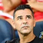 Míchel Sánchez, entrenador de fútbol, sentado en el banquillo con el escudo del Villarreal CF visible en el asiento, observando el campo con expresión analítica.
