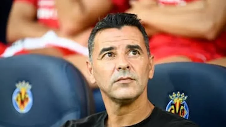 Míchel Sánchez, entrenador de fútbol, sentado en el banquillo con el escudo del Villarreal CF visible en el asiento, observando el campo con expresión analítica.