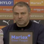 Primer plano de Hansi Flick durante una rueda de prensa, con expresión seria y analítica frente a micrófonos y un panel de patrocinadores de LaLiga y el Girona de fondo.