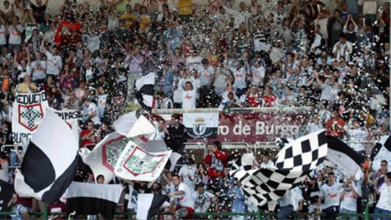 Aficionados en las gradas de un estadio de fútbol apoyando al equipo local con bufandas y banderas.