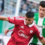 Jugador del Sevilla con camiseta roja disputa un balón con un defensor del Betis con camiseta verde y blanca durante el derbi de La Cartuja en LaLiga