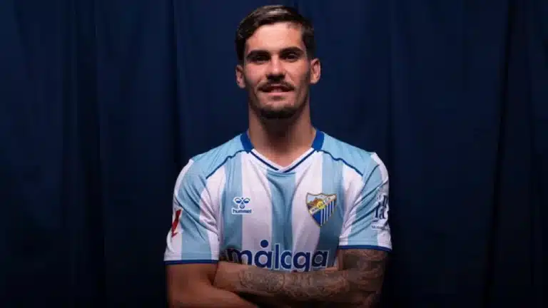 Chupete posa con los brazos cruzados y la camiseta blanquiazul del Málaga CF sobre un fondo azul oscuro