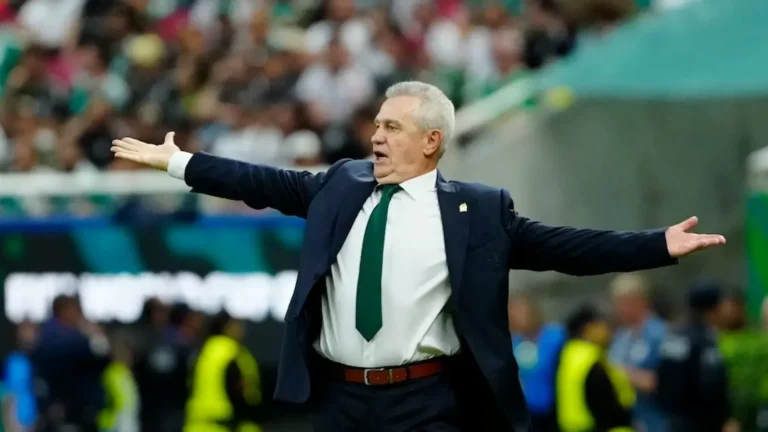 Javier Aguirre gesticula con los brazos abiertos en la banda, con traje azul oscuro y corbata verde