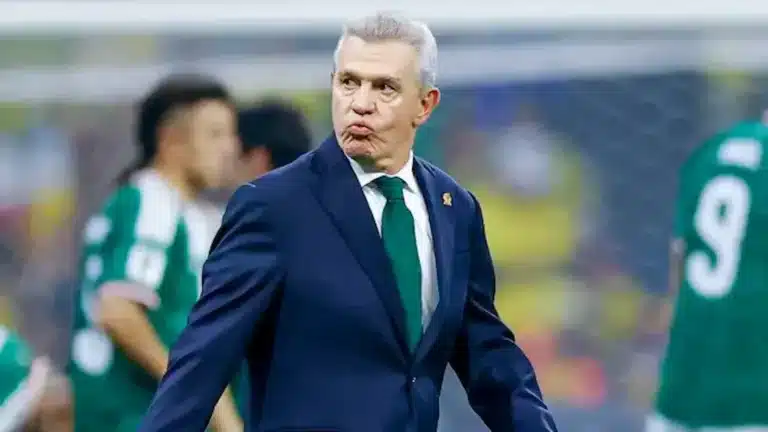 Javier Aguirre, con traje azul marino y corbata verde, gesticula durante un partido con jugadores de México al fondo