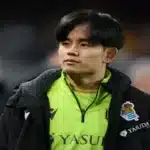 Take Kubo, con la chaqueta de la Real Sociedad sobre la camiseta verde, mira hacia un lateral del campo